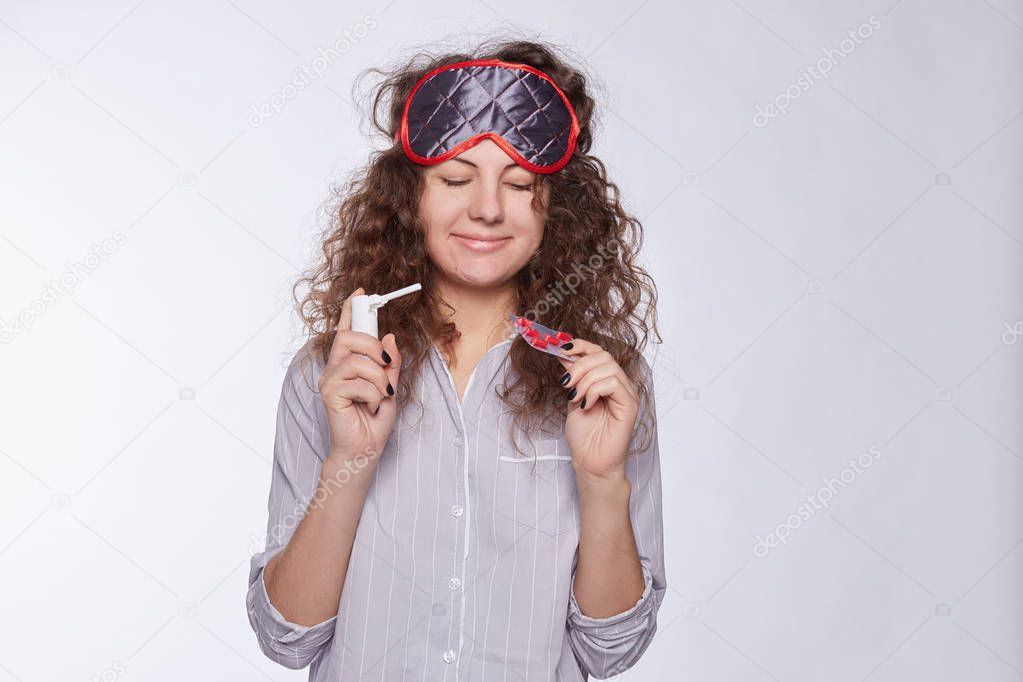 Cute glad Caucasian female is happy to get pills and medicine, ready to start treatment. Pretty ill young woman spent all day in bed indoors, wearing pajamas and sleeping mask, isolated on white wall.