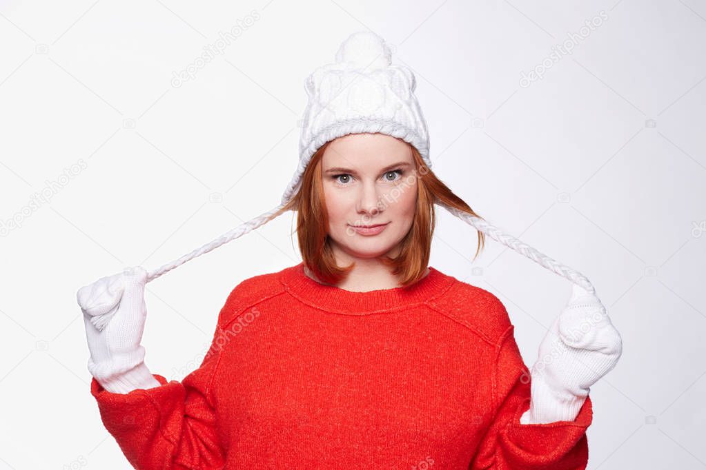 Red headed playful female, shows her two cute knitted pigtails on hat, has tender face expression, dressed in red sweater, isolated on white studio wall. Beautiful young woman having fun alone indoors