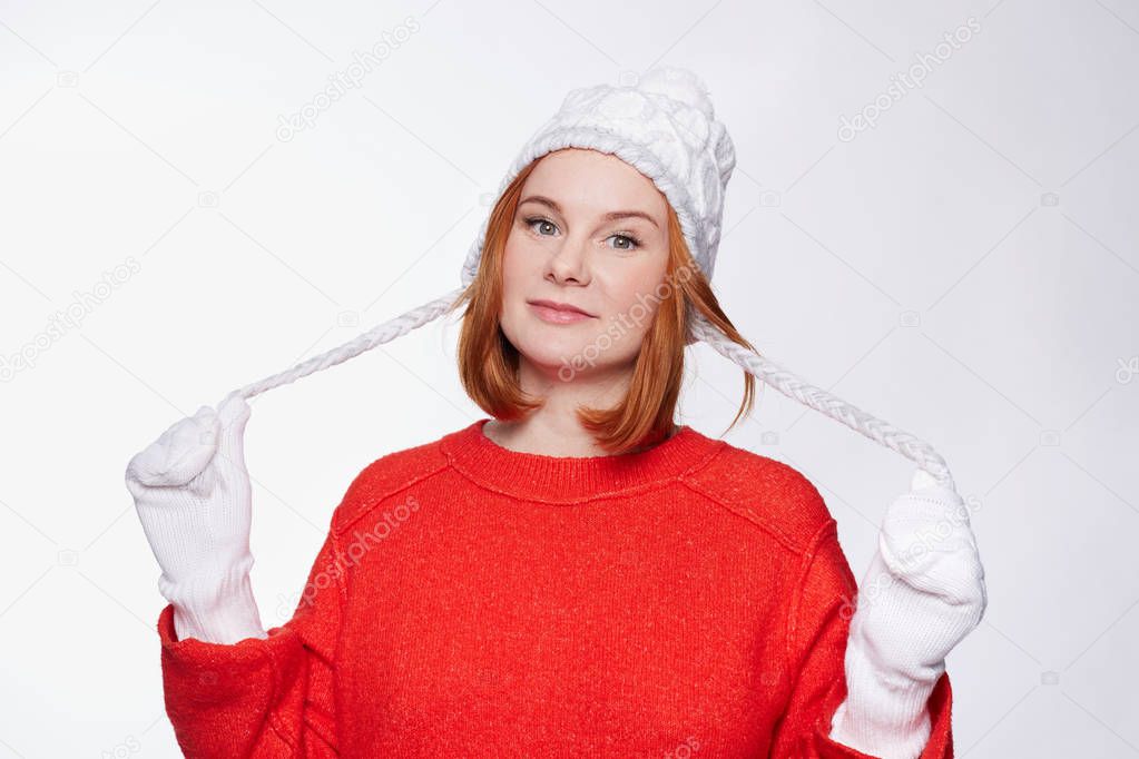 Waist up portrait of cute serious freckled female touches plaits from her knitted winter hat, dressed in red sweater and white mittens, posing in studio. Pretty woman model is going for a walk.