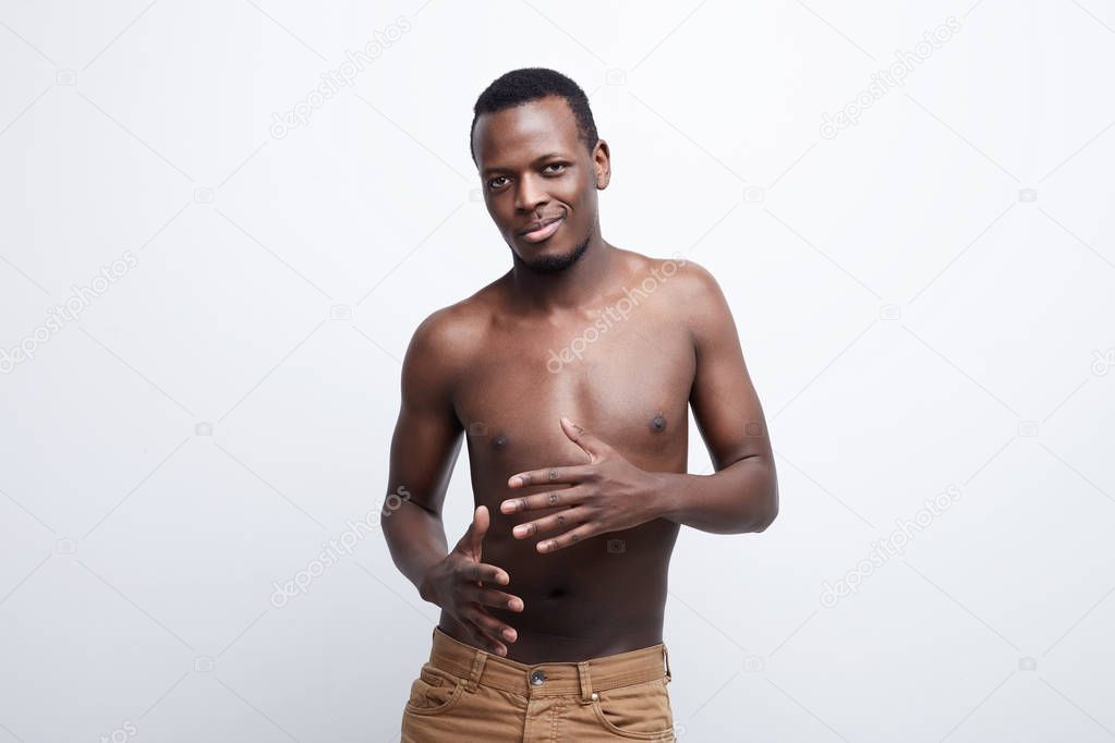 Handsome African American man with perfect shaped body is dancing, moving slowly, looking directly at the camera very serious. Seductive dark skinned posing shirtless , isolated over gray studio wall.