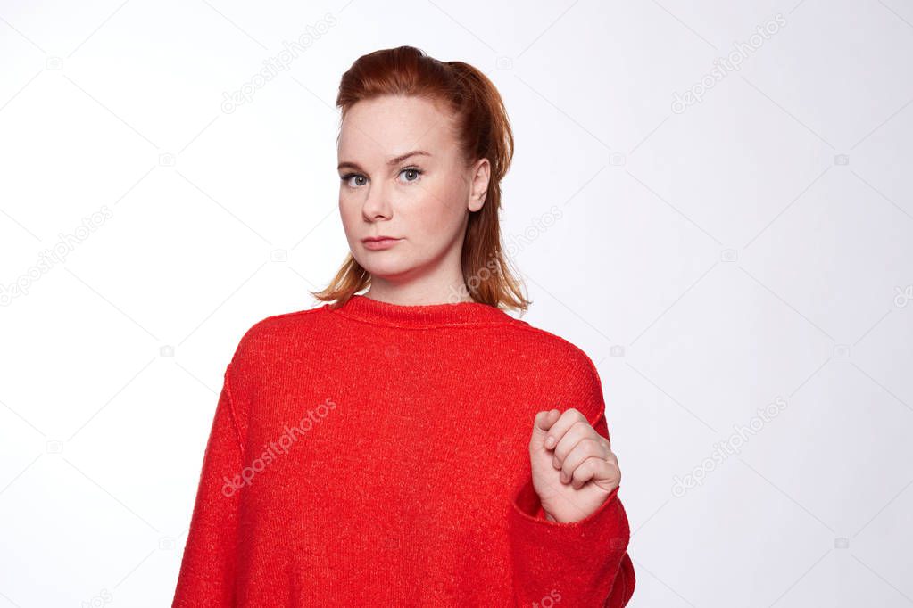 Elegant and serious Caucasian lady with freckles and red hairstyle looks with sensual glance at camera, having suspicious face expression. Woman does not believe her husband, afraid of being cheated.