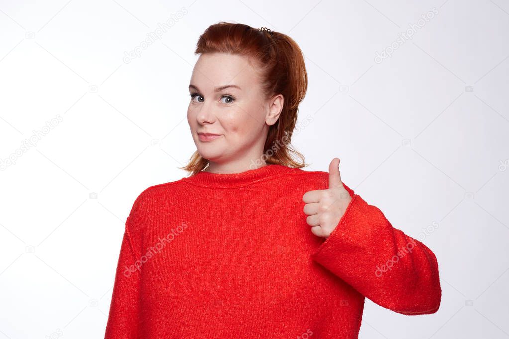 Close up of happy teenager keeps thumb raised, being in good mood, shows her agreement, poses over white background, says: Nice joke! Young woman shows like gesture, satisfied with choice you made.