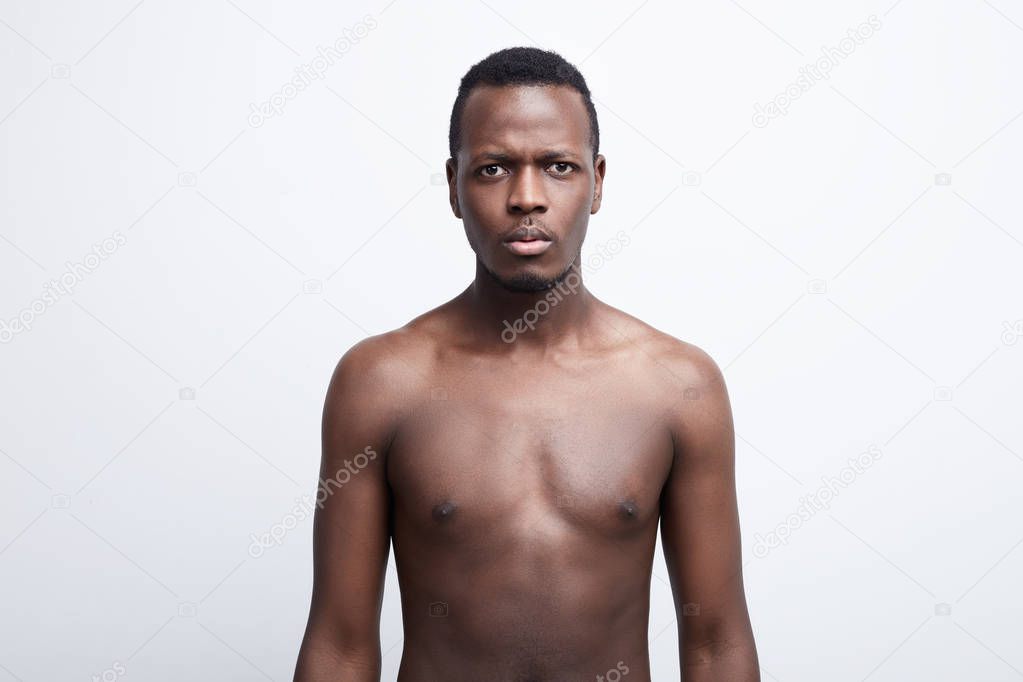 Close up shot of serious dark skinned male with scared and anxious emotion on face, feels astonishment as sees something unexpected, posing shirtless, isolated over white background. Human expressions