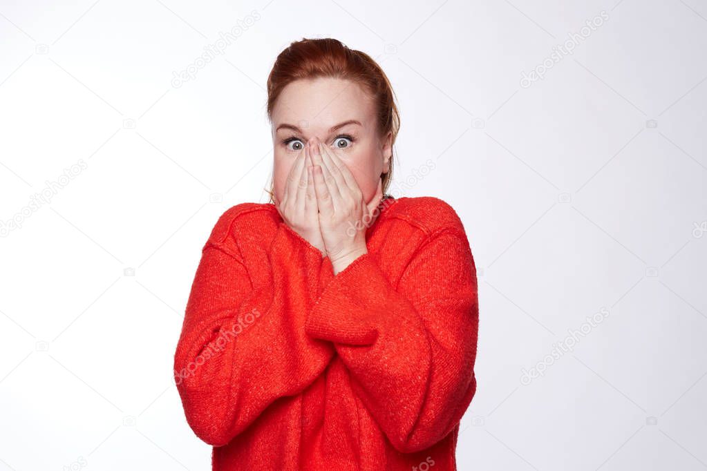 Astonished attractive funny woman with red hair covers face with both hands, does not want to spread rumours, shocked to hear sudden news from interlocutor, dressed in red oversized casual sweater.