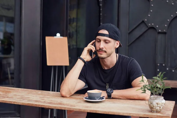 Attractive young European writer in black t-shirt, sitting at cafe with dreaming face expression, looking through the air, gaining inspiration in urban landscape around, mock up restaurant logo behind