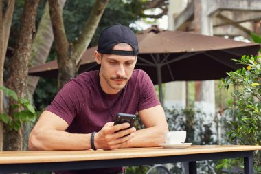 Kahve café'de sınıfları sonra üniversitede iken ciddi ve yoğun yüz ifadesi ile ekrana bakarak yakışıklı Avrupa öğrenci rahat t-shirt ve şapka cep telefonunuzun.
