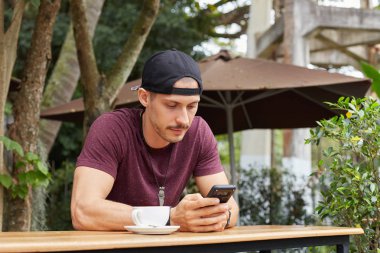 Kahve açık hava café'de kullanma online ücretsiz wi-fi, onun kız arkadaşı mesajlaşma şapka, moda aksesuarları yavaşça akıllı telefon, sms okurken gülümseyen yakışıklı genç Amerikalı erkek turist.