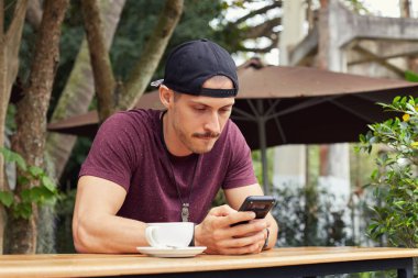 Kafede oturan iş üzerinde konsantre olmaya çalışırken alt dudak ısırma mobil uygulama içinde notlar yazma telefon, tutarak rahat sportif kıyafetler yakışıklı adam. Üniversite öğrencisi hazırlık ile meşgul