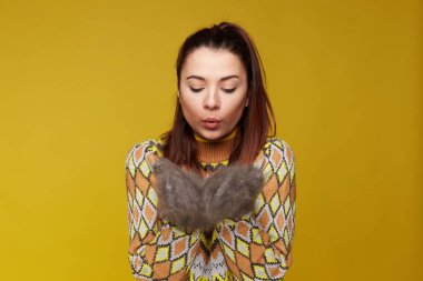Kuyruk uzun koyu saçlı çekici kadın, şık örme kıyafeti giymiş, doğal tavşan kürk onun sıcak eldivenleri üfleme, rahat ve sıcak duygu, sarı stüdyo duvar üzerinde izole.