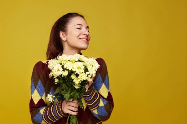 Yakın kadar genç beyaz kadının saçları at kuyruğu içinde geniş gülümsüyor, gözünden kapatma zevk, çiçek sevgili kişi var, sarı stüdyo modellerde duvar arka plan. Bahar tatil kavramı.