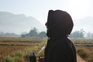 Outdoor portrait of brutal handsome adult man dressed in black jacket with hood is looking afar holding thermo cup, breathing fresh morning air in mountains, drinking hot beverage, thinking, resting.