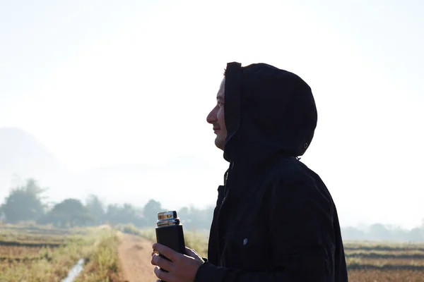 Profile close up portrait of male traveller dressed in black jacket with hood, holding thermos bottle with hot tea, looking around, breathing fresh morning air in mountains, enjoying nature alone.