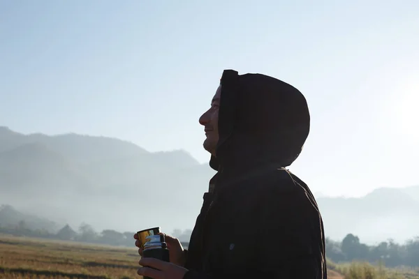 Profile close up portrait of male traveller dressed in black jacket with hood, holding thermos bottle with hot tea, looking around, breathing fresh morning air in mountains, enjoying nature alone.