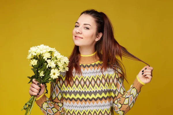 Caucasian coquette woman with flowers in hand, touching hair in pony tail, smiling gently, looking at camera, has pleased face expression, being in good mood. People, fashion, beauty, style concept.