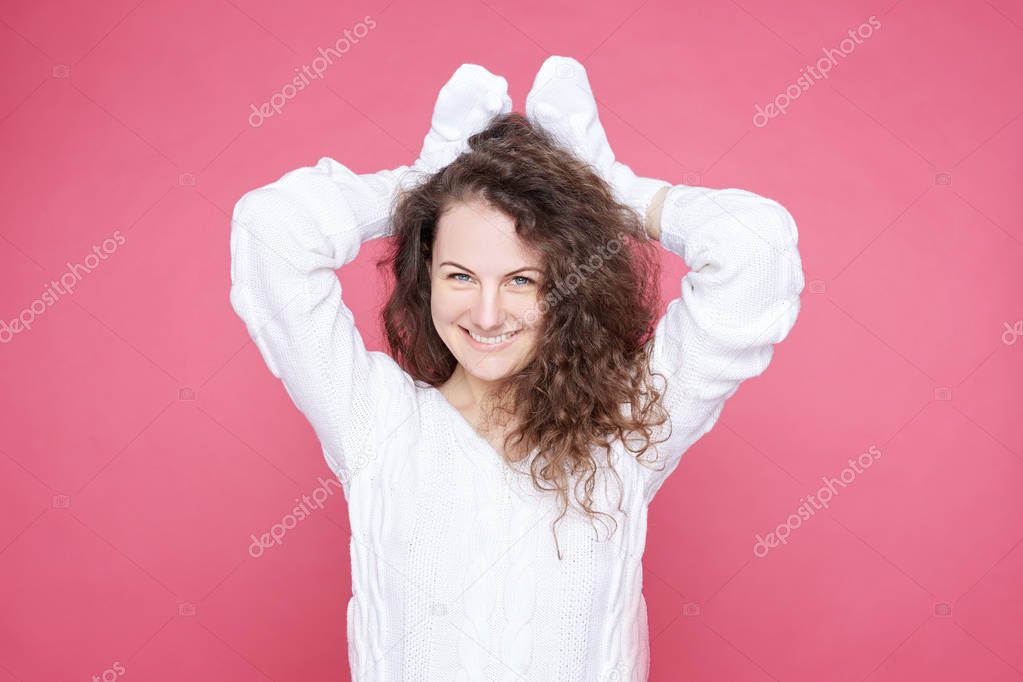 Indoor shot of cheerful woman holds hands over head as ears, imitates friendly adorable bunny, smiles broadly, wears knitted cozy sweater, plays with little child, stands indoor on pink background.