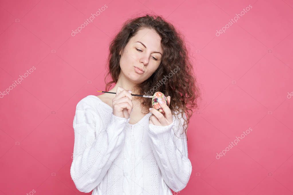  Concentrated curly haired lady holds traditional egg in hand, brush in other, painting it in different colors, rejoices coming holiday, wears cozy knitted outfit. Easter and spring celebration concept