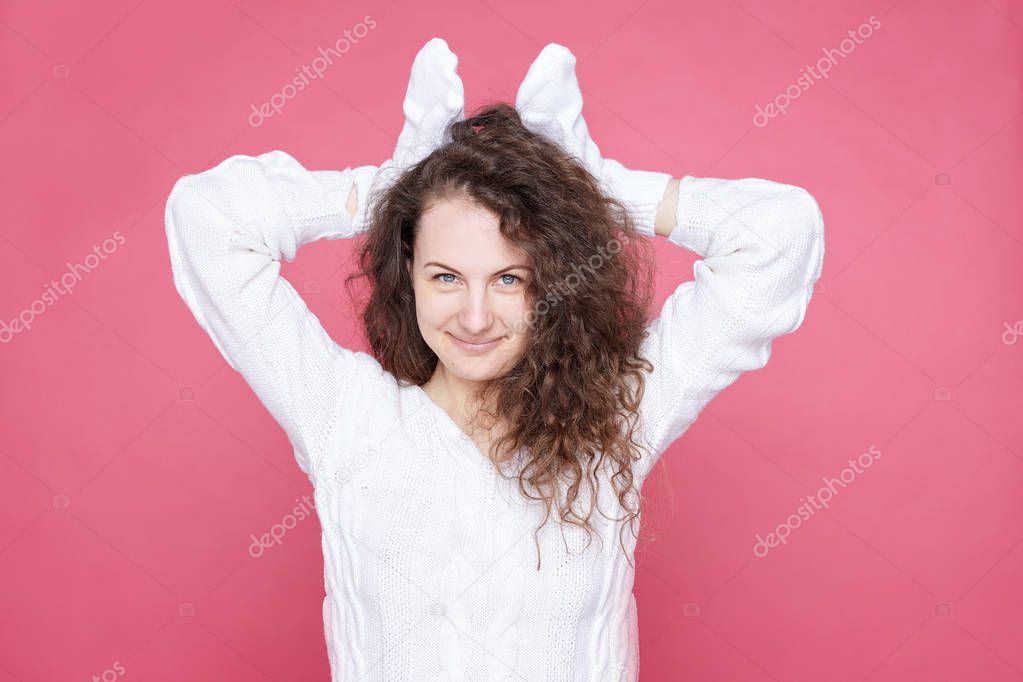 Close up shot of cute blue eyed curly haired lady holds hands in white mittens over head as ears, imitates friendly adorable bunny, having fun with family, smiles broadly, ready to celebrate Easter.