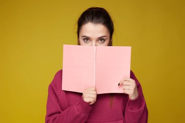 Stresli utangaç kadın öğrenci cevap korkuyor, pembe kitap arkasında gizler, Lila hoodie giyer, farklı kaynaklardan malzeme öğrenir, sarı arka plan üzerinde izole, kamera konsantre görünüyor.