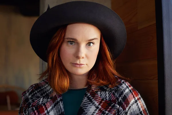 Close up of ginger haired freckled attractive romantic woman in black hat, sitting in modern cafe, having serious face expression. Elegant Caucasian lady looking at camera while having rest indoors.