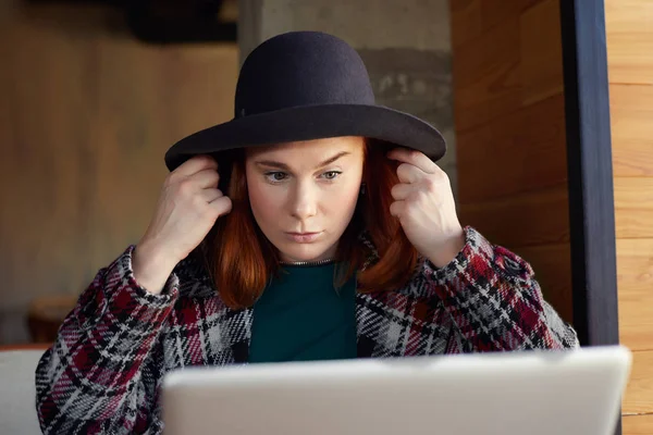 People and work concept. Indoor shot of concentrated beautiful woman dressed in black hat and coat, sitting at wooden desk,  using modern device for work and as mirror, waiting for boyfriend to come.