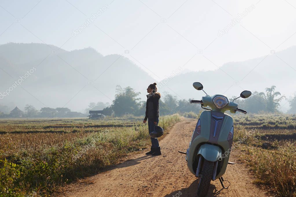 Stylish female traveller leaving motorbike to look around picturesque nature with helmet in hand, admiring fields and mountains, feeling happy and inspired. Woman with hair bun had long way before.