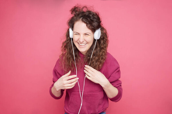 Positive Caucasian woman with curly haircut, keeps both hands on headphones, listens favorite song, connected to unrecognizable device, feels boost of happiness from good sound. Isolated on pink wall.