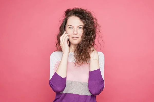 Emotional blue eyed female speaks via cell phone, tells news to friend, dressed in fashionable summer colorful outfit, looks at camera with wonder, stands on pink, copy space aside. Lifestyle concept.