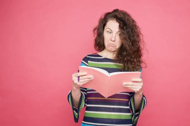 Şaşkın sorgulanan beyaz öğrenci, gazetelerde şüpheyle ifade değil bakar, pembe defter taşır, cevap bulmaya çalışır, rahat renkli gömlek giymiş, pembe duvara izole.