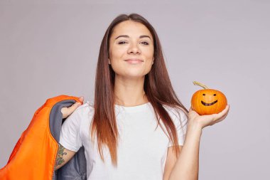 Pleased calm mixed raced young woman harvesting pumpkin, tilts head, smiles broadly, holds orange jacket, enjoys autumn time and holidays, isolated. Traditional Halloween symbol in lady's hands. clipart