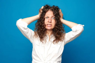 Depresyonda olan Avrupalı bir kadın kafasını kaşıyor, hayal kırıklığına uğramış bir yüz ifadesiyle bakıyor, yanlış yaptığı için pişman, beyaz gömlek giyiyor, mavi arka planda poz veriyor. İnsanlar, olumsuz duygular ve duygular.