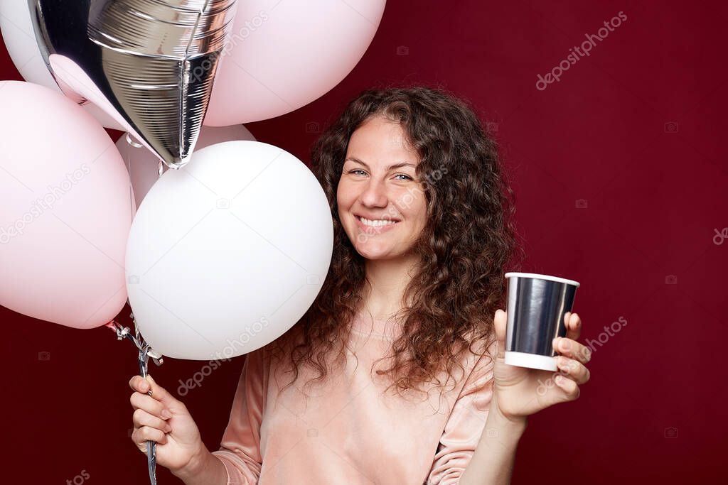 Young female in casual clothes being on birthday party, looking at camera with perfect toothy smile and holding colorful balloons in hand. Cheerful woman celebrating  anniversary on crimson studio wal