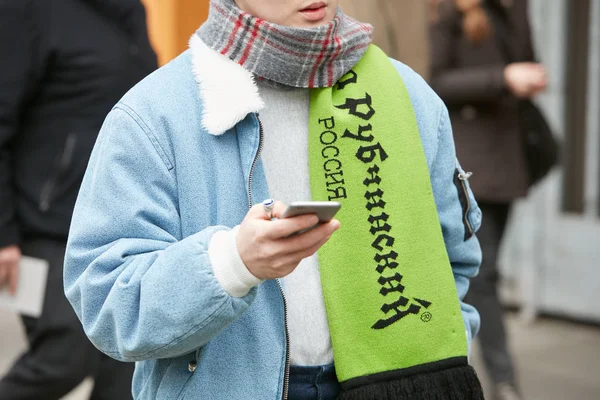 Мужчина в зеленом шарфе и синей джинсовой куртке смотрит на смартфон перед показом мод Джорджио Армани, уличный стиль Недели Моды в Милане 15 января 2018 года . — стоковое фото