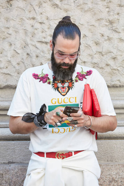 Мужчина в белой футболке Гуччи и красной сумке, проверяющий смартфон перед показом мод Сальваторе Феррагамо, уличный стиль Недели моды в Милане 18 июня 2017 года
.