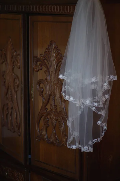 White tulle wedding veil with pretty detailed embroidered flowers around the front rim, perched on a vintage dark brown ornate wardrobe door, in preparation for wedding ceremony for the bride