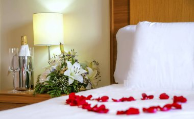 Gül yaprakları ile odak komidinin üstünde sıcak ve romantik yatak. Yatak takımları, çiçekler, şampanya flüt beyaz, küme yukarı balayı