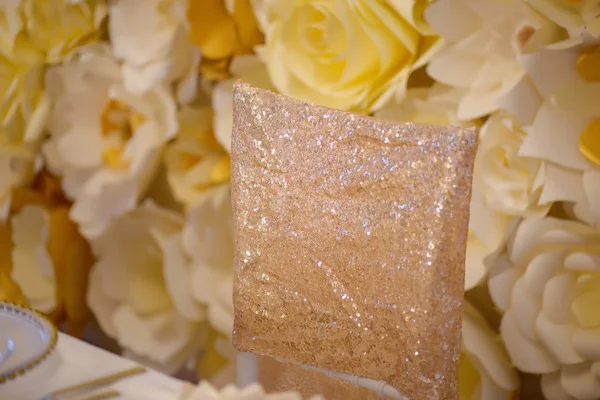 Sitting arrangement at a formal event or fine dining restaurant featuring transparent plates with golden details, glassware and silverware in the order of use set against a paper flowers wall