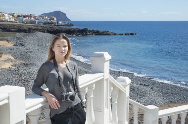 Kafkas kökenli genç bir kadın boynunda dijital, profesyonel bir kamera tutuyor, çalışıyor ya da araştırıyor, okyanusla önden görüşlü ve arka planda küçük bir çakıl taşı plajı var.