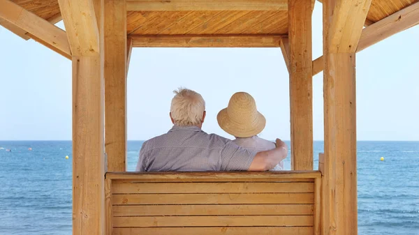 Senior çift, şapka ve beyaz gümüş saçlı adam ile sahil şeridinde bir ahşap gölgelik altında oturan ve el Medano, Tenerife, Kanarya Adaları, Ispanya romantik bir kucaklamak okyanusa bakarak kadın