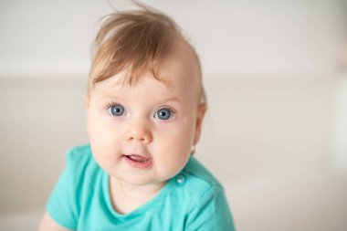 Mavi gözlü sevimli beyaz kız çocuğunun portresi, sakince kameraya bakıyor, merakla, etkileşimle veya merakla beyaz bir bebek karyolasında oturuyor; şirin bebek ifadeleri