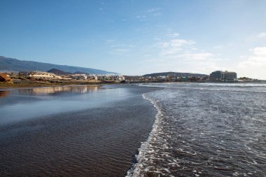 Güzel geniş ve sakin volkanik plaj, Playa de la Jaquita, kıyı yerleşim yerlerine ve otellere doğru kısmi iç manzara, ve yakınlardaki kaya oluşumları, El Medano, Tenerife, Kanarya Adaları, İspanya