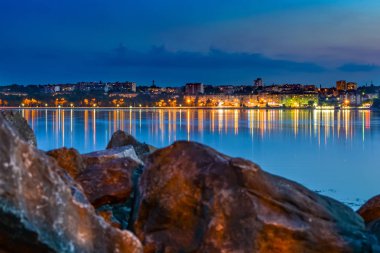 Doğu Avrupa şehrin ışıkları göle yansıtılır. Taş bloklar ön planda.