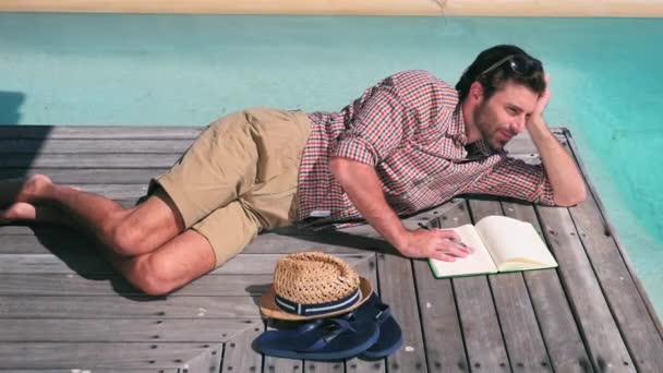 プールのそばの木の床の上に横たわるノートブックと若い男 — ストック動画