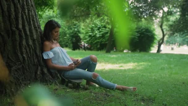 女性は木に対してスケートボードに座って スマートフォンを使用して — ストック動画