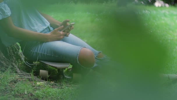 女人坐在滑板上靠着树 用智能手机 — 图库视频影像