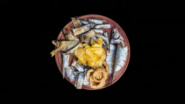 Набор закусок для пива. Соленая, сушеная рыба, кольца кальмара и картофельные чипсы, вращайтесь на разделочной доске. Изолированный на черном фоне . — стоковое видео