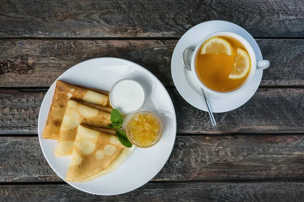 Deliciosas panquecas com geléia e creme azedo no prato e xícara de chá com limão . — Fotografia de Stock