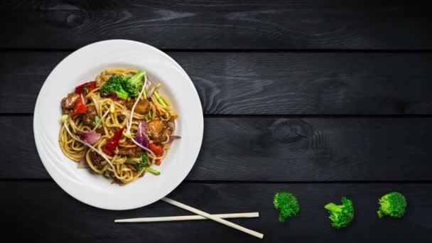 Περιστρεφόμενο αναδεύεται τηγανητά noodles udon με κρέας ή κοτόπουλο και λαχανικά. Διακοσμημένα με φρέσκο μπρόκολο και chopsticks σε μια λευκή πλάκα σε μαύρο φόντο ξύλινη. Το top view με το χώρο αντίγραφο για το κείμενό σας — Αρχείο Βίντεο