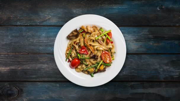 Nouilles à frire udon rotatives avec fruits de mer dans une assiette blanche sur fond en bois. Vue du dessus — Video