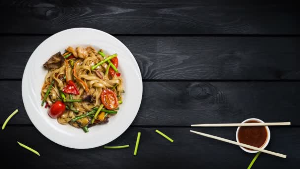 Εκ περιτροπής ανακατώνει τα τηγανητά noodles udon με θαλασσινά και λαχανικά. Διακοσμημένα με φρέσκα φασόλια τα λάχανα και chopsticks σε μια λευκή πλάκα σε μαύρο φόντο ξύλινη. Το top view με το χώρο αντίγραφο για το κείμενό σας — Αρχείο Βίντεο