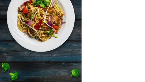 Roterande udon stir fry nudlar med kött eller kyckling och grönsaker. Dekorerad med färsk broccoli i en vit platta på trä bakgrund. Fyrkantig layout för sociala nätverk och kopia utrymme för din text — Stockvideo